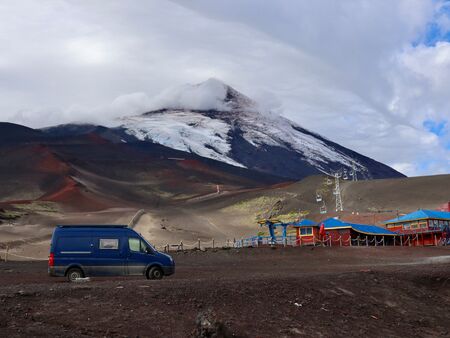 Ein blauer Van steht in einer fantastischen Vulkanlandschaft auf schwarzer abgekühlter Lava. Im Hintergrund der teils Schneebedeckte Osorno Vulkan. Rechts neben ihm ein bunter Sessellift. Das Wetter ist bewölkt. Das Bild trotzdem beeindruckend.
