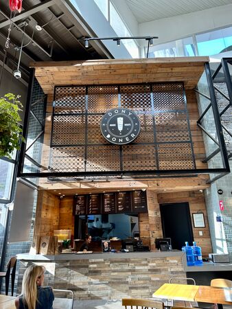 Moderner Döner-Kebab-Stand „Dony Doner“ in einem Einkaufszentrum mit Holz- und Metalldesign. Eine Frau arbeitet hinter der Theke, während eine blonde Kundin im Vordergrund sitzt. Natürliches Licht strömt durch die Deckenfenster.