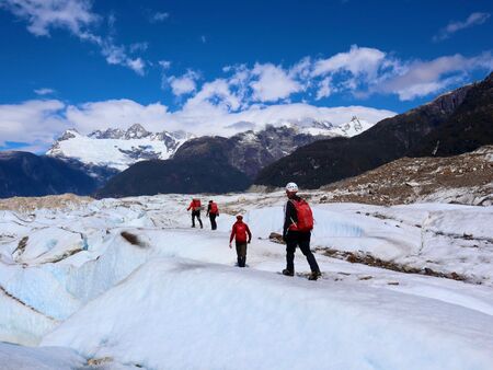 4 Erwachsene mit Steigeisen wandern mit roten Rucksäcken und Helm auf dem Glaciar Exploradores in Chile. Im Hintergrund schneebedeckte Berggipfel, der Himmel strahlt blau und überall ist Gletschereis. Ein abenteuerlicher Anblick der zum Erkunden einlädt.