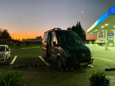 Ein blauer Van steht auf einem Parkplatz an einer Autobahn-Tankstelle. Im Hintergrund geht die Sonne unter. Auch das ist Vanlife… manchmal muss eben eine Tankstelle herhalten.