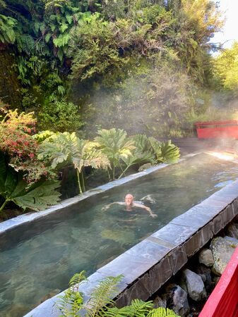 Ein Mann schwimmt in einem sauber und vor Wärme dampfenden Termalbecken. Wunderschön angelegt inmitten von üppiger grünen Natur und roten Holzstegen, die durch die Beckenlandschaft führen. Er sieht sehr glücklich und entspannt aus.