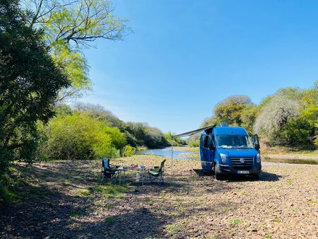 Blauer VW Crafter Camper an einem idyllischen Fluss in Uruguay mit ausgefahrener Markise.