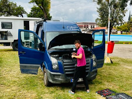 Ein Mann in einem pinken T-Shirt arbeitet an einem blauen Van mit geöffneten Türen und Motorhaube auf einem Campingplatz in Salta, Argentinien. Im Hintergrund sind weitere Fahrzeuge und Gebäude zu sehen.