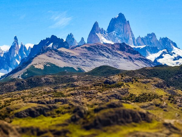 Eine atemberaubende Landschaft, vom Mirador de las Águilas aus fotografiert, die den Fitz Roy und den Cerro Torre bei klarem Wetter zeigt. Untermalt wird das Foto von satten Farben wie grün, blau und ein leichtes grau-violett, welches die Bergkette betont