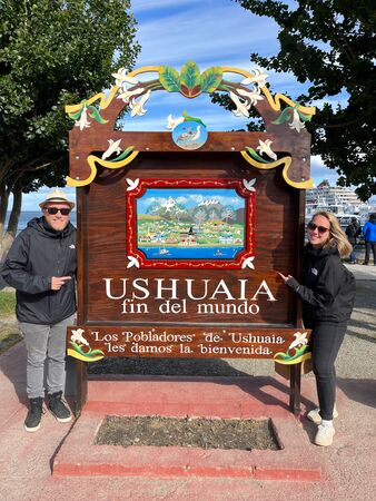 Zwei Personen stehen lächelnd neben einem kunstvoll gestalteten Schild in Ushuaia, das als “Fin del Mundo” (Ende der Welt) bekannt ist. Der Hintergrund zeigt einen malerischen Hafen und einen klaren blauen Himmel.