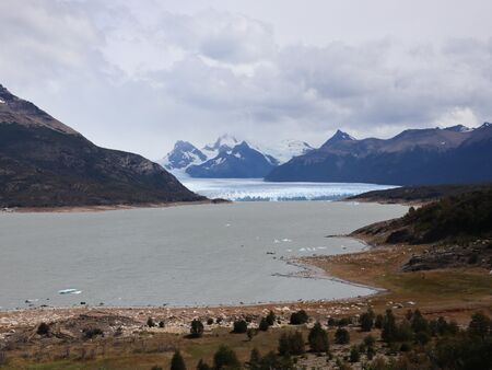 Ein weiter Blick auf den Perito Moreno Gletscher in Patagonien, Argentinien, umgeben von Bergen und einem See im Vordergrund, bei bewölktem Himmel.