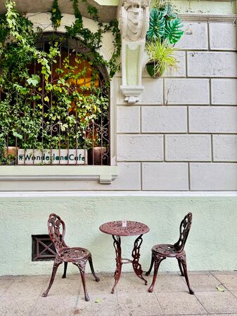 Eine idyllische Café-Ecke im Freien im Barrio Lastarria, Santiago de Chile, mit einem kleinen Metalltisch und zwei passenden Stühlen. Die Szene ist von üppigem Grün umgeben, das über ein dekoratives Eisengitter rankt.