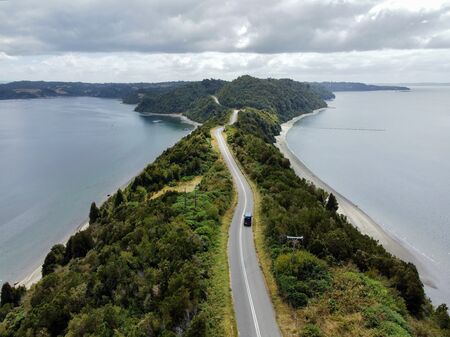 Eine Drohnenaufnahme von einem blauen Van, der auf einer landschaftlich wunderschönen Landzunge fährt. Rechts und links befinden sich grüne Büsche und Bäume, bevor anschließend Sandstrand und Meer auf beiden Seiten gleichermaßen kommen. Es ist bewölkt.