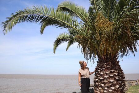 Eine Frau steht neben einer Palme an der Aussichtsplattform der Bastion de San Pedro in Colonia del Sacramento, Uruguay, mit dem Rio de la Plata im Hintergrund. Die Szene zeigt die natürliche Schönheit und historische Bedeutung dieses malerischen Ortes.