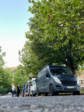 Drei Vans stehen am Straßenrand einer asphaltierten Straße. Mehrere junge Erwachsene stehen gesellig zusammen. Ein Vanlifer-Treffen.