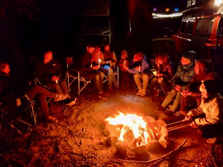 9 Erwachsene und 2 Kinder sitzen in einem großen Halbkreis um ein orangerot leuchtendes Lagerfeuer herum. Niemand blickt in die Kamera, sondern alle unterhalten sich oder sind mit Marshmellows grillen beschäftigt. Es sieht sehr einladend aus.