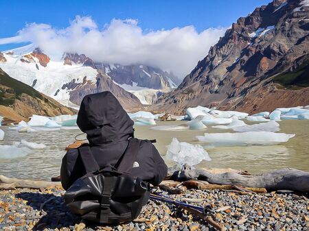Der Rücken eines sitzenden Mannes mit Kapuze auf vor einem See (Laguna Torre in Patagonien) voller Gletscher-Eis-Stücke.