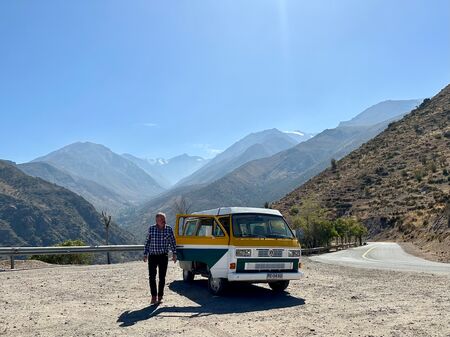 Das Foto zeigt eine Person, die neben einem farbenfrohen, gelb-grün-weißen VW T3-Bus auf einer Bergstraße steht. Im Hintergrund erstrecken sich dramatische, karge Bergketten unter einem strahlend blauen Himmel.