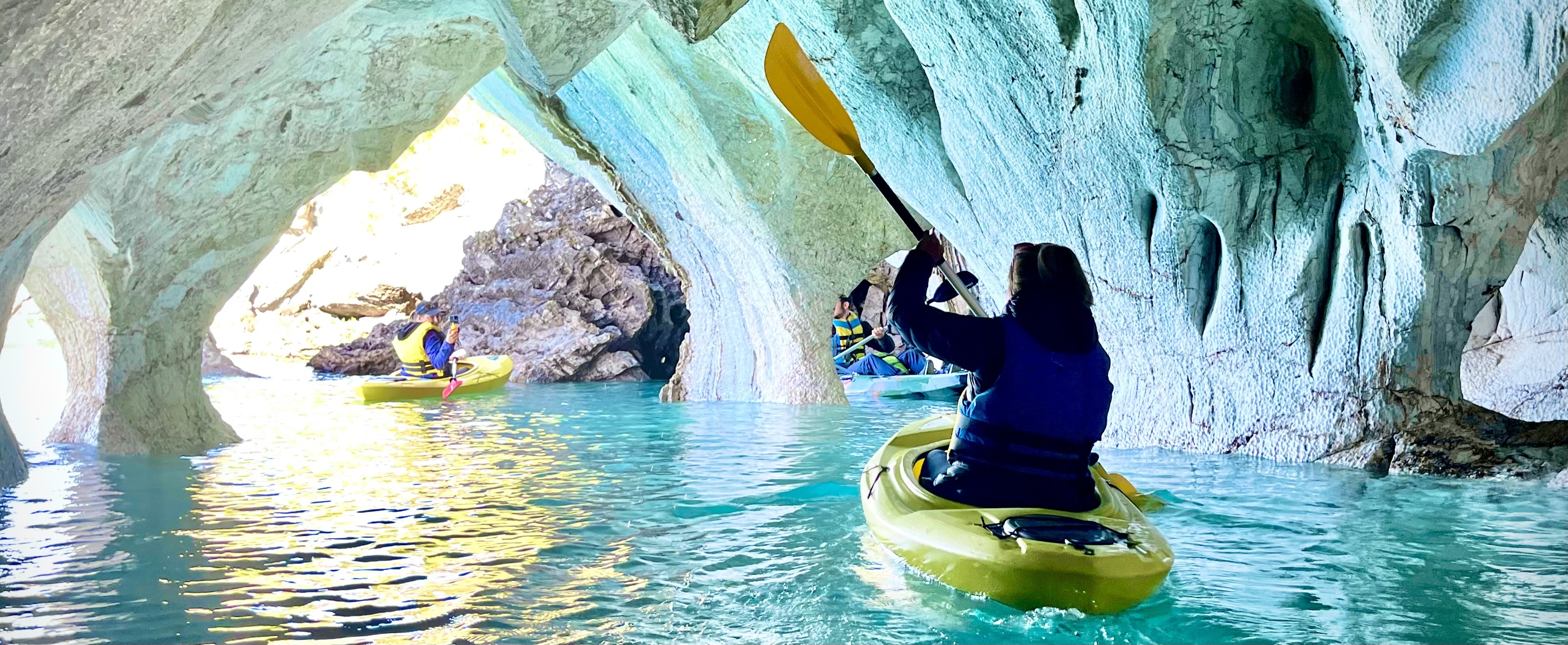 Eine Kajakfahrerin in einem gelben Kajak auf türkisblauem Wasser. Sie fährt durch eine Höhle - die Catedral de Marmol in Chile auf der Carretera Austral. im Hintergrund zwei weitere Kajakfahrer. Die Höhle reflektiert das Blau des Wassers. Wunderschön.